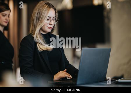 Professional woman works on laptop in a modern office, focused and confident, with colleague nearby Stock Photo