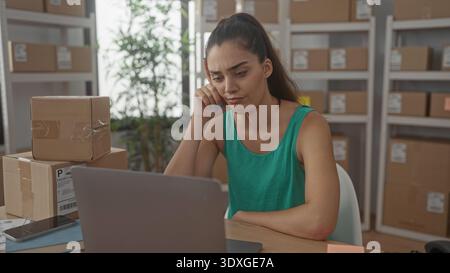 Woman rests hand on cheek scrolling on laptop beside stacked packages in building; ecommerce shipping anxiety. Stock Photo