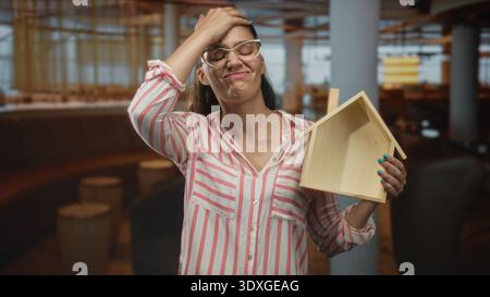 Woman wearing striped shirt and glasses holds wooden house model with hand on forehead in restaurant; frustration. Stock Photo