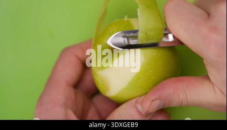 Hands peeling a green apple with a knife on a bright green cutting board during a kitchen task Stock Photo