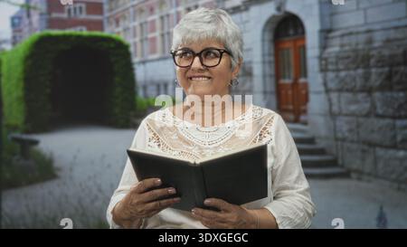 Woman with grey hair holding a black book, hands visible, reading by a stone building entrance in a courtyard; serenity reflection. Stock Photo