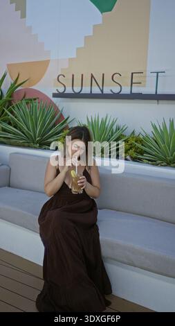 Woman sipping iced tea through straw while holding glass on a building terrace with succulents and sign reading sunset; serenity. Stock Photo