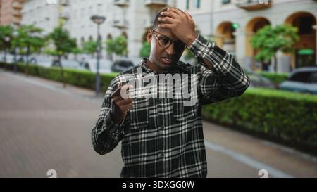 Young man holding car keys with hand to forehead on a city street near parked cars and hedges, wearing glasses and plaid shirt; frustration. Stock Photo