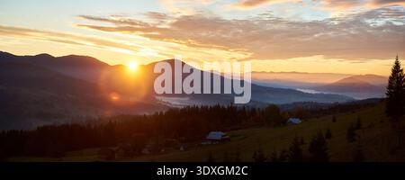 Sun rises over rolling hills, casting warm golden glow across landscape. Soft mist blankets valley, colors decorate trees. Silhouetted pine trees and few scattered homes add depth morning scene. Stock Photo