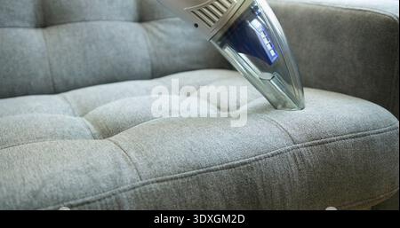 Vacuum cleaner removes dust from a gray couch in a bright living room during the day Stock Photo
