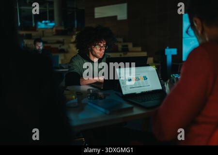 Focused coder with glasses works on laptop in a dim office, showcasing collaboration and concentration Stock Photo