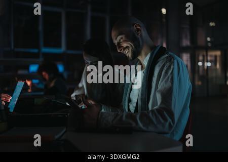 Smiling man using tablet in a dim coworking space with colleagues nearby, conveying focus and teamwork Stock Photo
