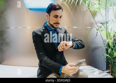 Professional businessman checks time on smart watch while using smartphone in a modern office lobby Stock Photo