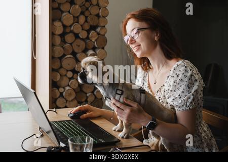 A smiling redhead woman in glasses works on her laptop with a small cute dog on her lap. Concept of remote work, pet-friendly office, and modern Stock Photo