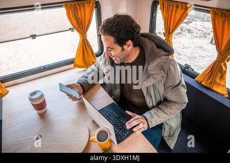 Man smiling, using a smartphone and laptop at a table inside a camper van, enjoying the digital nomad lifestyle Stock Photo