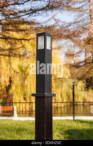 Modern black metal bollard lamp on green grass in a public park. The blurred background features warm yellow autumn foliage, creating a peaceful, sere Stock Photo