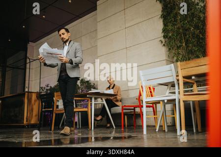 Professional man in a gray blazer reads papers at an outdoor cafe, with a woman working nearby Stock Photo