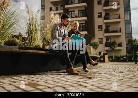 Business partners review documents on a bench outside a modern building, discussing plans together Stock Photo