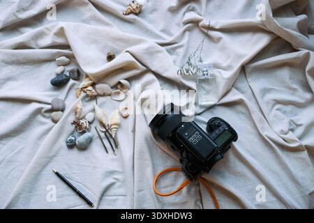 Digital camera resting on fabric surrounded by assorted seashells, pebbles, pen and small glass perfume bottle, capturing still life arrangement with natural elements and technology Stock Photo