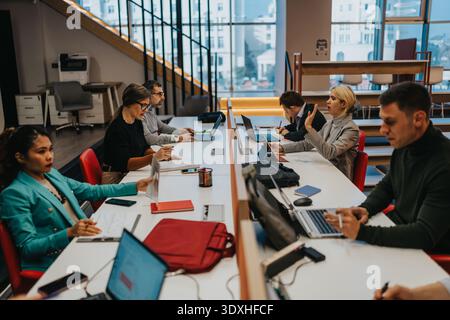 Diverse team of professionals collaborating at a long office desk with laptops and papers Stock Photo