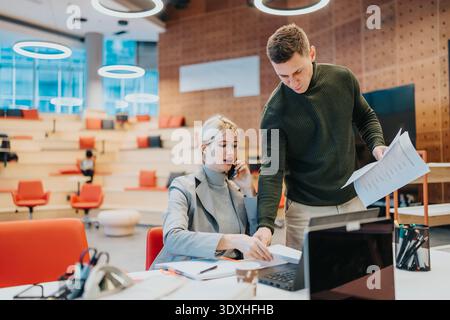 Two professionals collaborate in a modern office, reviewing documents and using a laptop together Stock Photo