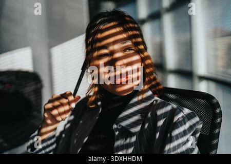 Professional woman in striped light shadows at a modern office, holding a pen Stock Photo