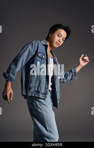 Young Asian man confidently poses in denim, showcasing style in a grey studio. Stock Photo