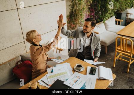 Two business partners celebrate with a high-five over documents in a modern cafe office. Stock Photo