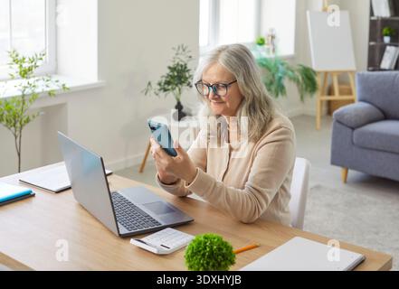 Senior woman holding mobile phone to call family or read news, sitting at home table with laptop Stock Photo
