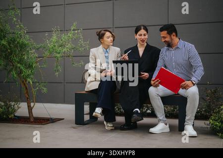 Three colleagues discussing documents and laptop while sitting on an outdoor bench during a business collaboration Stock Photo