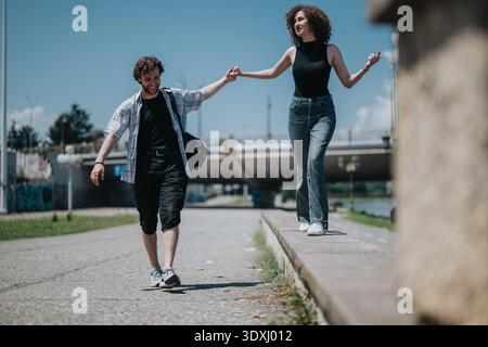 Friends walk hand in hand along a riverside path under a bridge on a sunny day Stock Photo