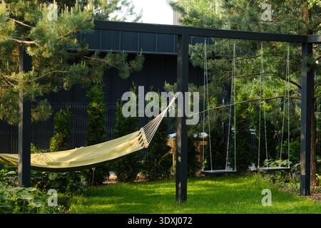 A hammock hangs between two posts in a garden surrounded by tall green trees. Two swings are nearby, with grass underneath. The sun shines down on thi Stock Photo