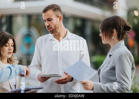 Three professionals discuss documents outdoors in an urban city setting, collaborating on a project Stock Photo