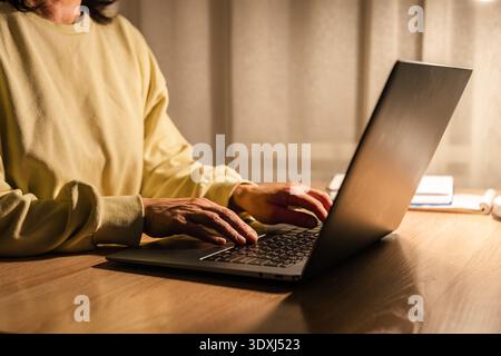 Woman working remotely on a laptop, under warm, soft lighting at a desk. Person typing on laptop keyboard at home at night Stock Photo