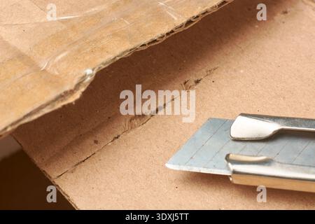 utility knife blade cutting brown cardboard box, sharp steel cutter on textured corrugated surface, closeup with copy space, unboxing shipping package Stock Photo