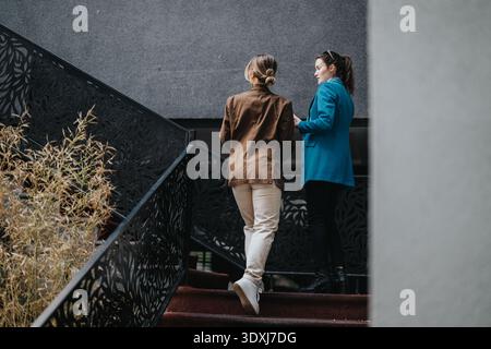 Two women in stylish jackets converse on an outdoor stairway in an urban setting Stock Photo