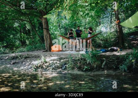 Friends relax in a hammock at a forest campsite by a river and trees Stock Photo
