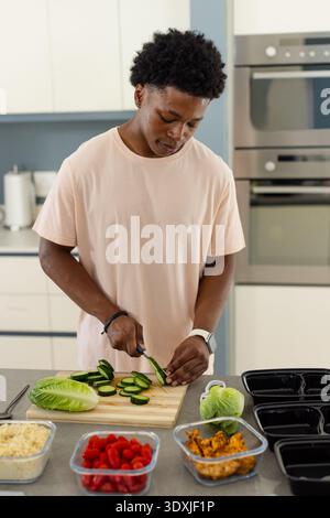 African American man in pink shirt slicing cucumber on board in kitchen assembling meal trays Stock Photo