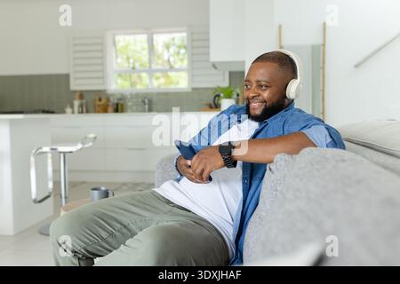 Man reclining on sofa at home holding smartphone, wearing headset and black smartwatch, smiling Stock Photo
