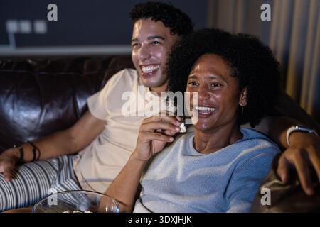 Diverse men in loungewear sitting on leather couch at home, watching TV, sharing popcorn bowl Stock Photo