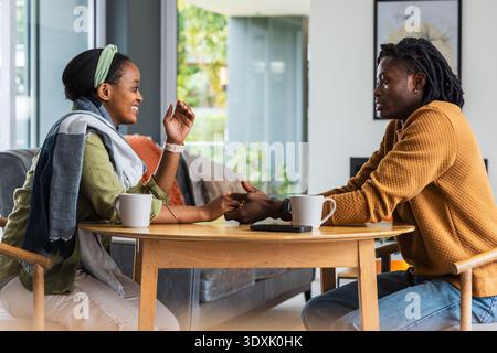 African American couple holding hands chatting at home table wearing scarf with two mugs and phone Stock Photo