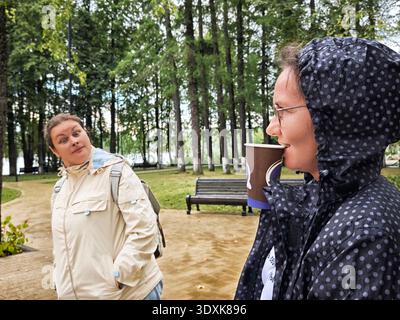 Middle aged woman enjoys coffee while chatting with friend in a green park during a rainy day Stock Photo