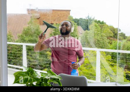 Adult African man cleaning glass sliding door on balcony, holding squeegee and spray bottle Stock Photo