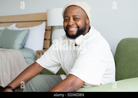 African American man sitting on green chair near bed with pillows, wearing beanie and headphones Stock Photo