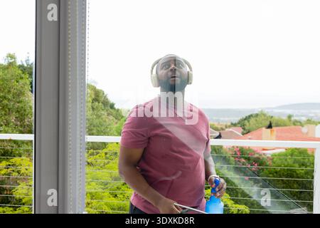 African man standing on balcony cleaning glass door with blue bottle squeegee wearing headphones Stock Photo