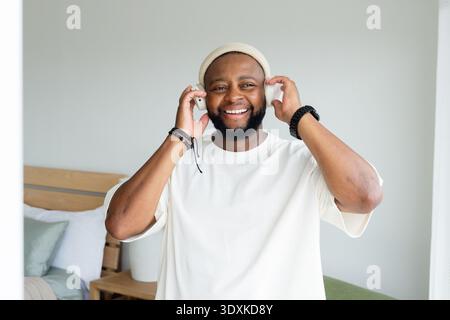 Man smiling and holding headphones while listening with smartphone in bright bedroom wearing beanie Stock Photo