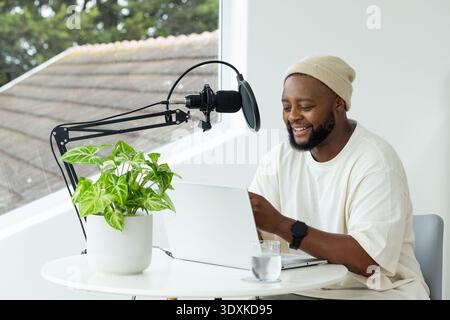 African man sitting at white table in bright studio, using laptop and microphone, wearing beanie Stock Photo
