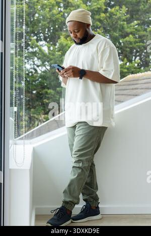 African American man standing at stair landing beside floor-to-ceiling window, checking smartphone Stock Photo