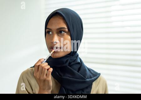 African woman applying lip gloss with wand near lips at home by window blinds wearing hijab Stock Photo