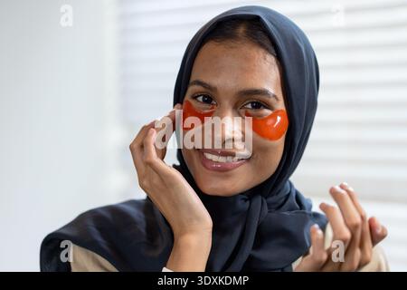 Woman wearing dark navy headscarf applying orange under-eye hydrogel patches by window blinds Stock Photo