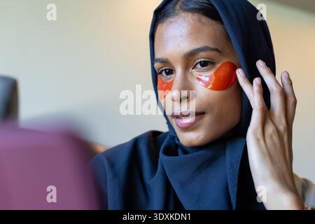 Woman wearing dark hijab touching orange gel under-eye patches sitting at vanity with pink device Stock Photo