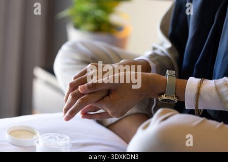 Woman applying cream, massaging hands on white bedspread with open jar and wristwatch Stock Photo