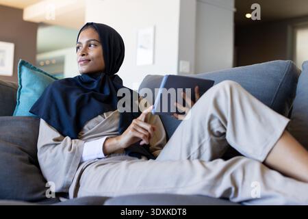 Woman reclining on sofa with teal pillow holding dark tablet tapping screen wearing navy headscarf Stock Photo