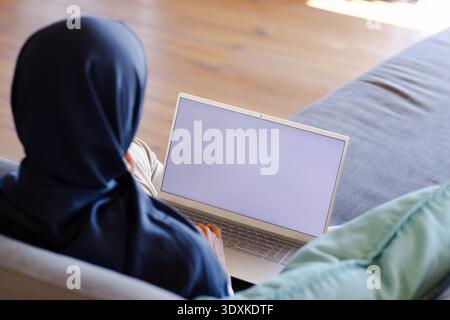 Adult woman wearing dark headscarf typing on silver laptop while sitting on grey sofa, copy space Stock Photo
