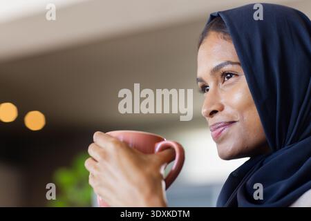 Woman wearing dark navy headscarf holding pink ceramic mug near mouth, smiling in cafe, copy space Stock Photo